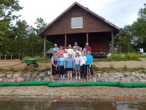 Almost all the people who volunteered to bring the beach project to completion are shown here on the workday at the edge of the lake. 1st row l-r: JoAnn Hopkins, Celeste Snitko, Sarah Sangermano, Margaret Blackburn, Gabrielle Oja and Rebecca Oja. 2nd row l-r: Janice Moore, Nancy Campbell, Greg Blackburn, Wendy White and Mary Shonk. Back row: Hank Campbell, Judy Knapp, Jeff Oja, Walter Snitko and Bronson Shonk. Missing from the photo were Connie Cerroni and Joe Sangermano. Photo by Emily Shonk Schoelzel